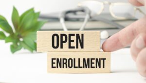 Close-up of wooden blocks with the words Open Enrollment next to a plant and eyeglasses: What Is Open Enrollment