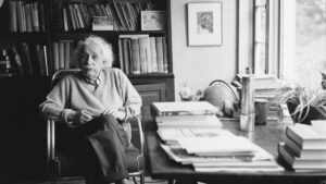 Black and white photo of a person sitting in a study surrounded by books and papers on a large desk.